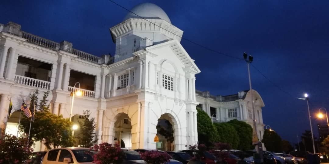 A large, white colonial-style building with a central dome and arched windows, surrounded by parked cars and street lamps.