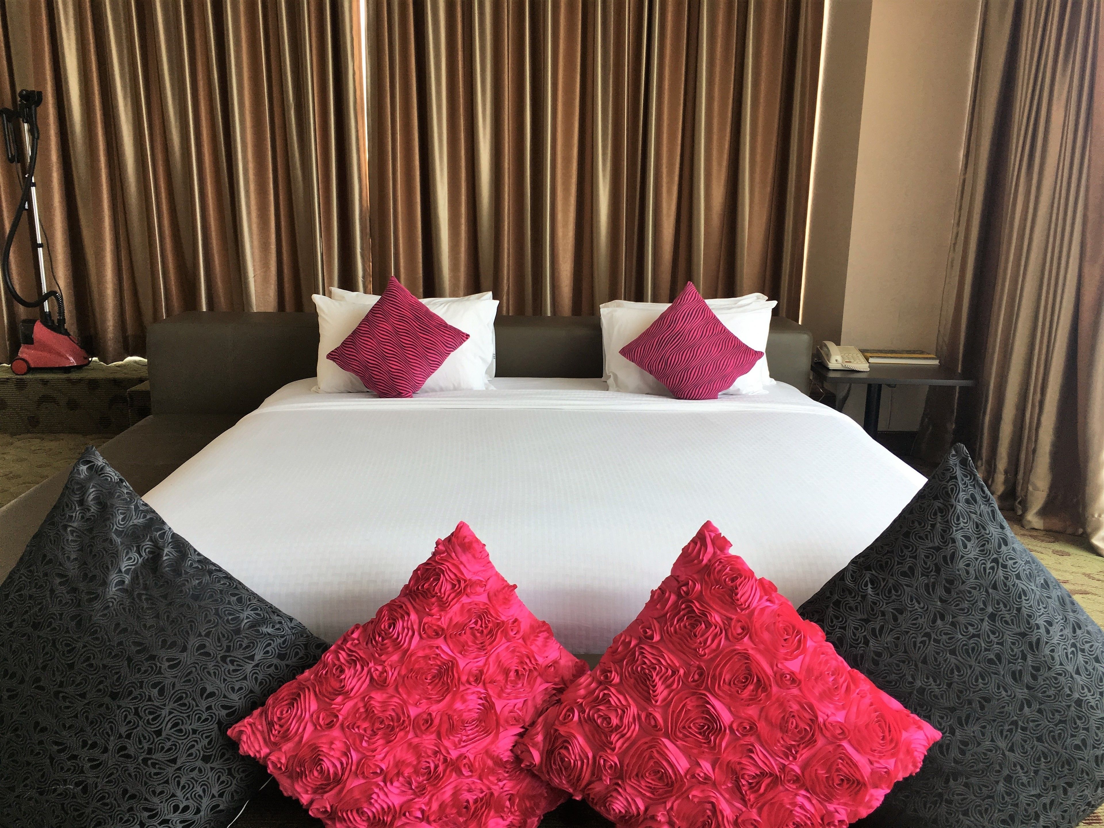 A hotel room with a large bed, two pillows, a bedside table, and a curtain backdrop.