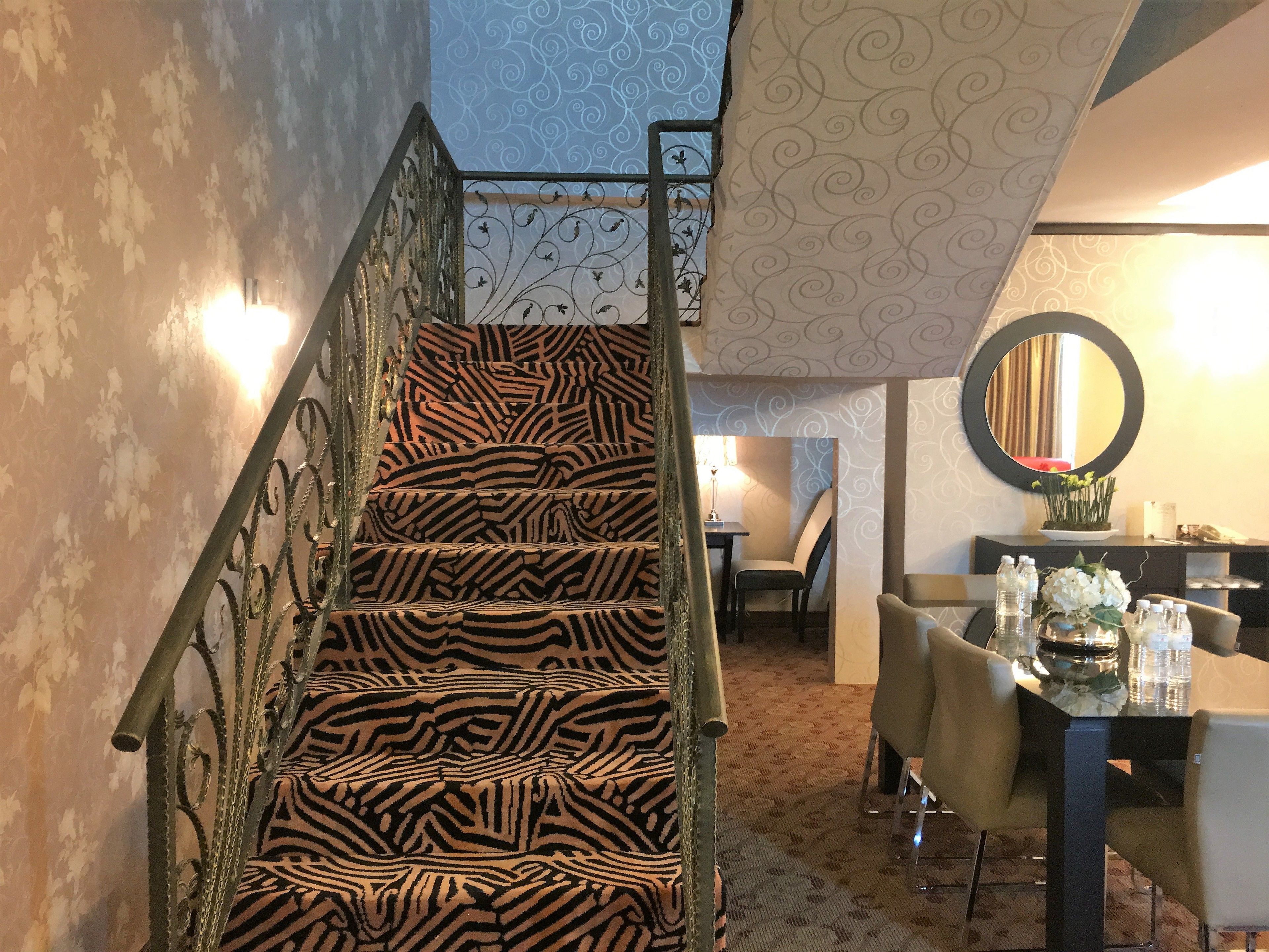 A staircase with zebra-striped carpet leading to a dining area with a table and chairs, a mirror on the wall, and a patterned wallpaper.