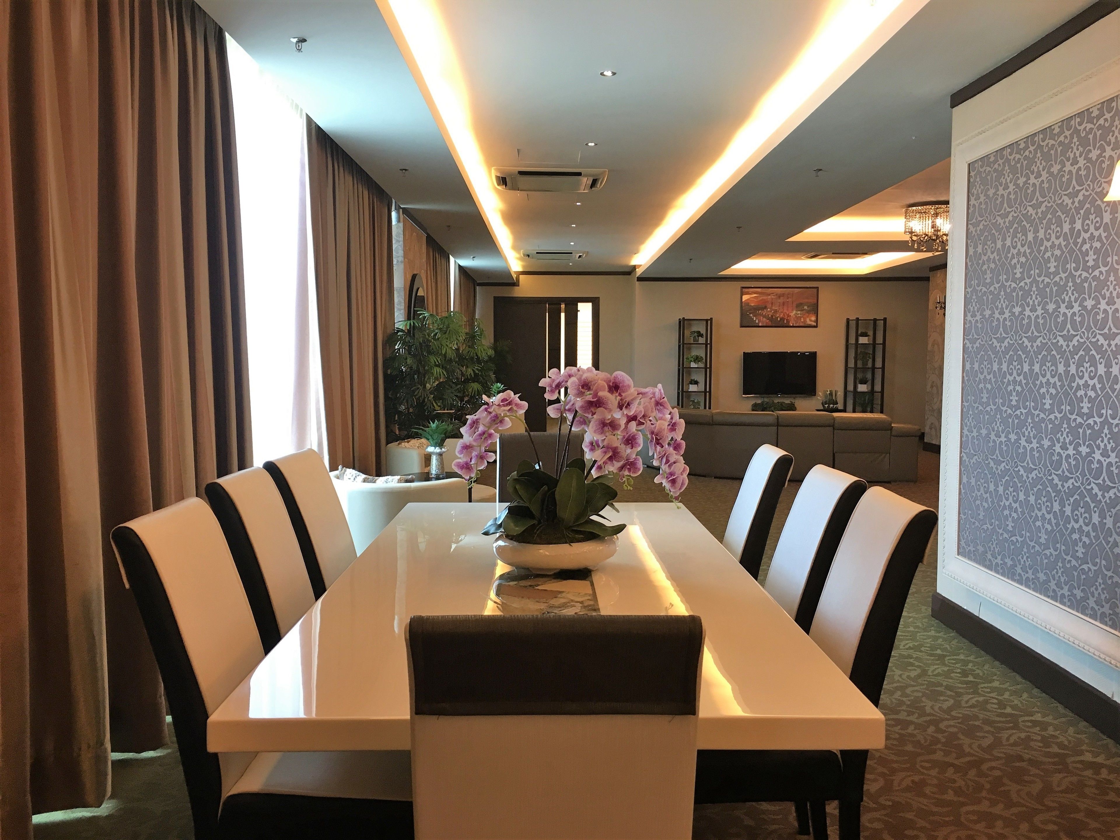 A hotel dining area with a long table, six chairs, a vase of flowers, and a television in the background.