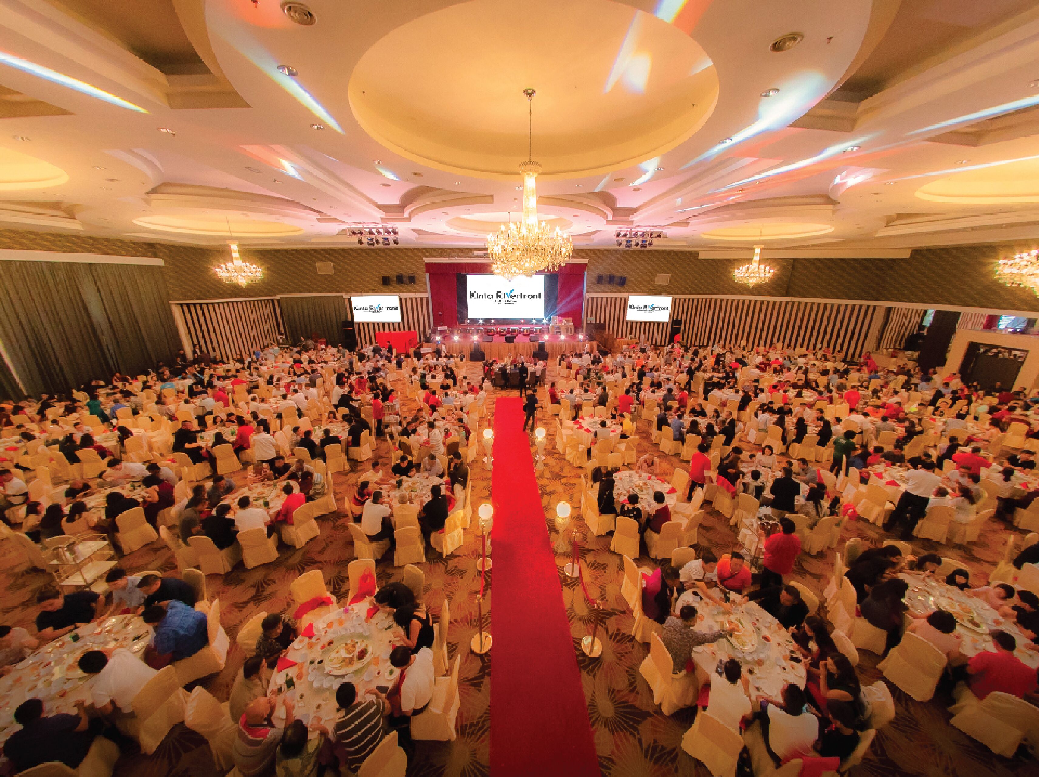 A large banquet hall with round tables and chairs, a central stage, and a chandelier.