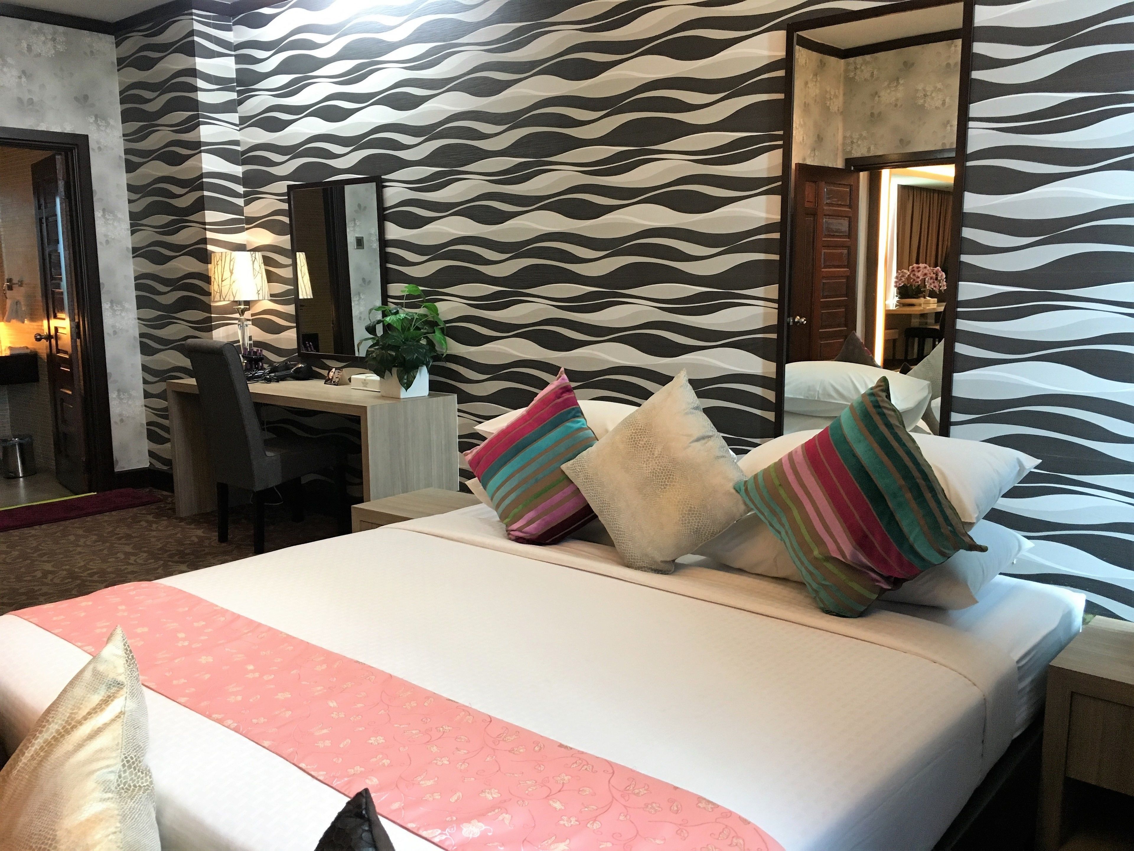 A hotel room with a bed, a desk, and a chair. There is a mirror on the wall and a plant on the desk.