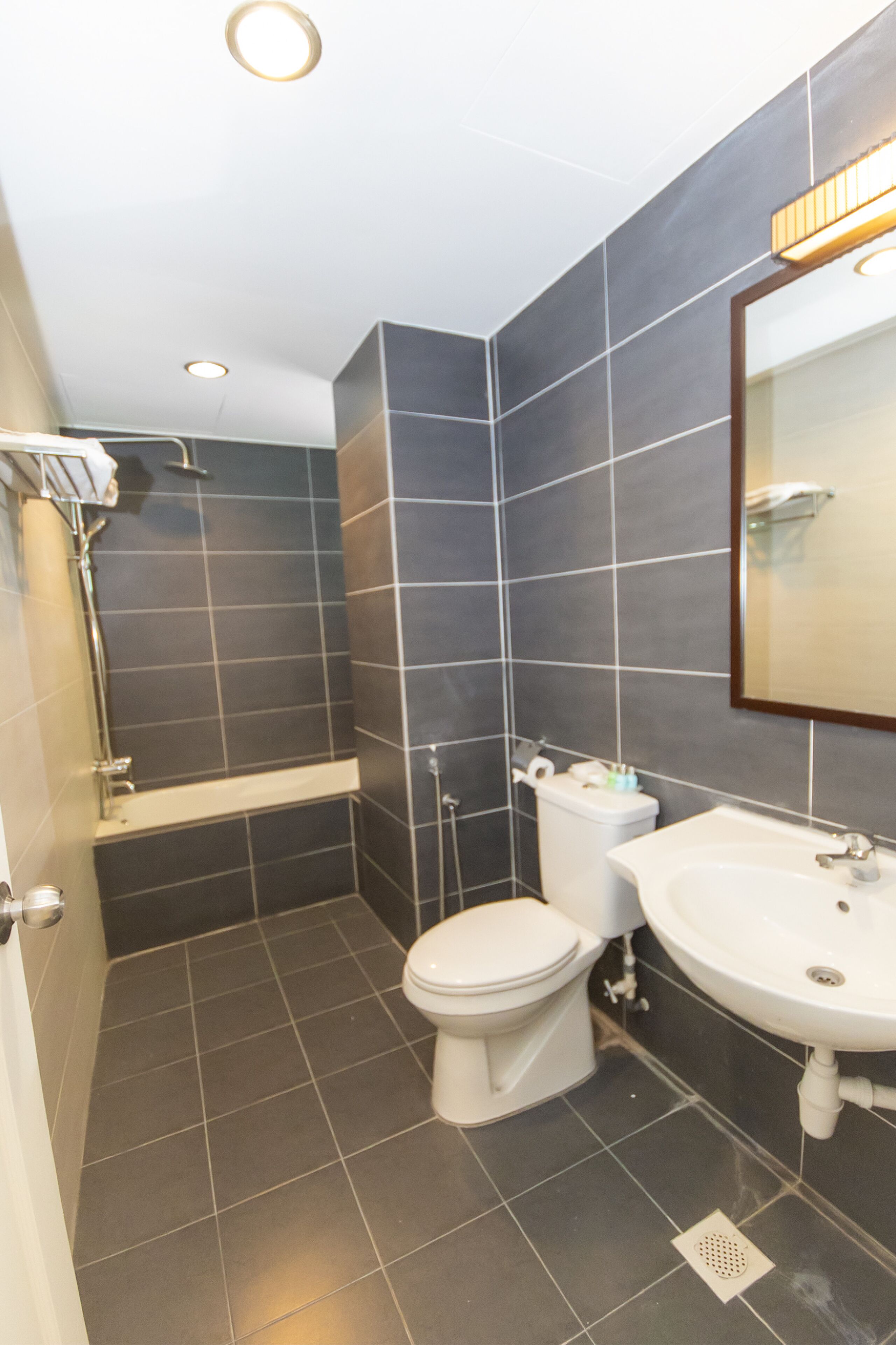 Un baño moderno con azulejos oscuros, un inodoro blanco, una bañera y un lavabo con espejo.