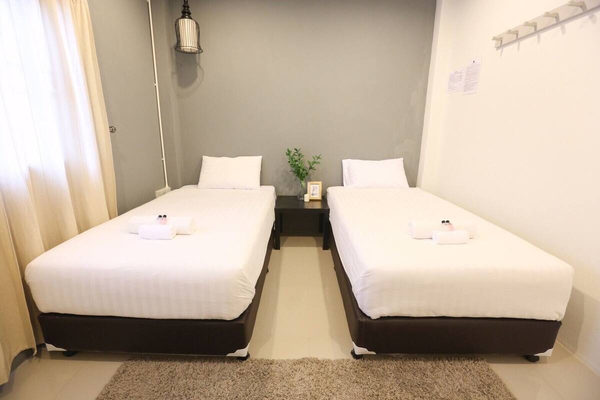 Ein Hotelzimmer mit zwei Einzelbetten, einem kleinen Nachttisch mit Lampe, einer Pflanze und einem Wandregal.