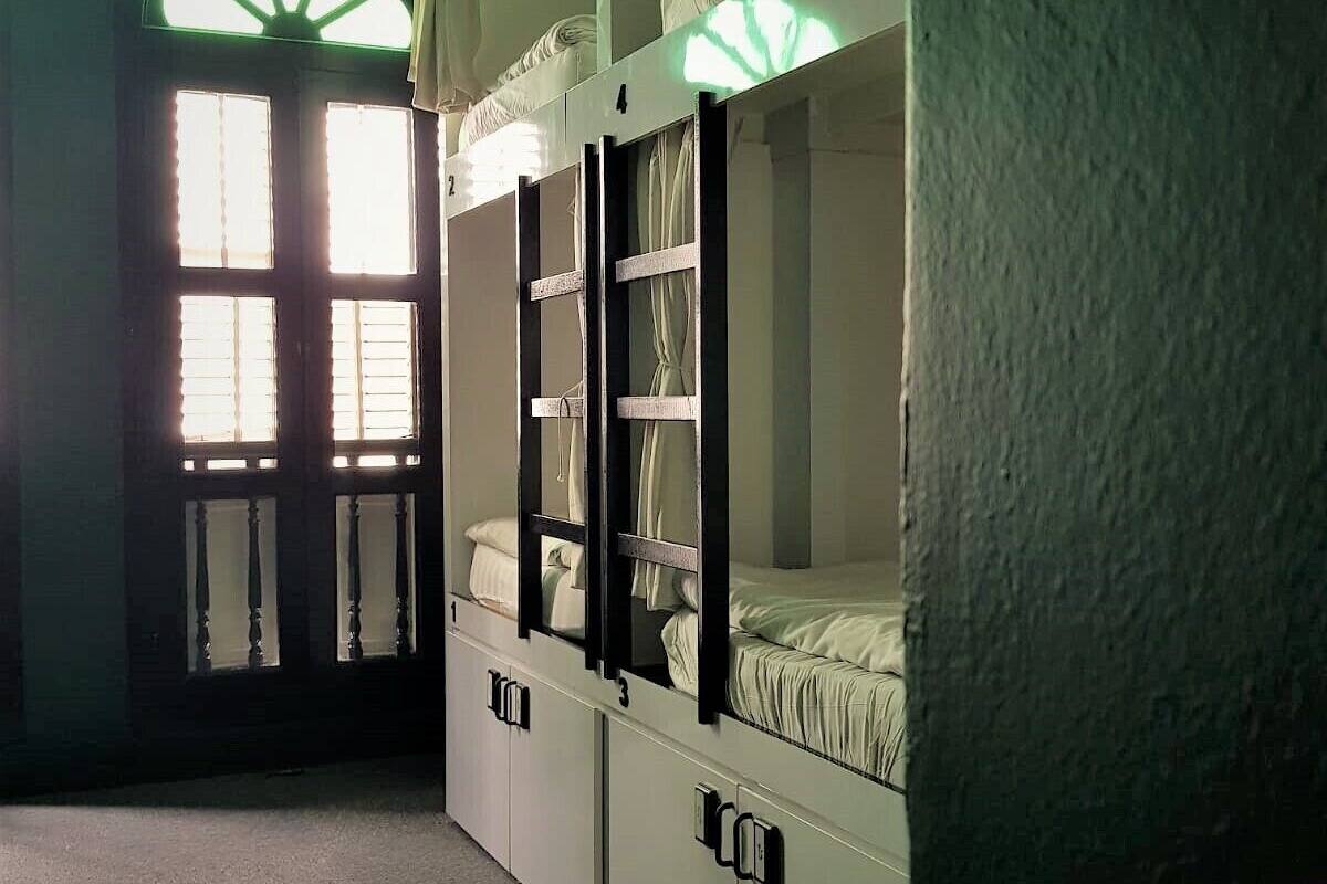 A dormitory room with bunk beds, a window with a green light, and a door with a number.