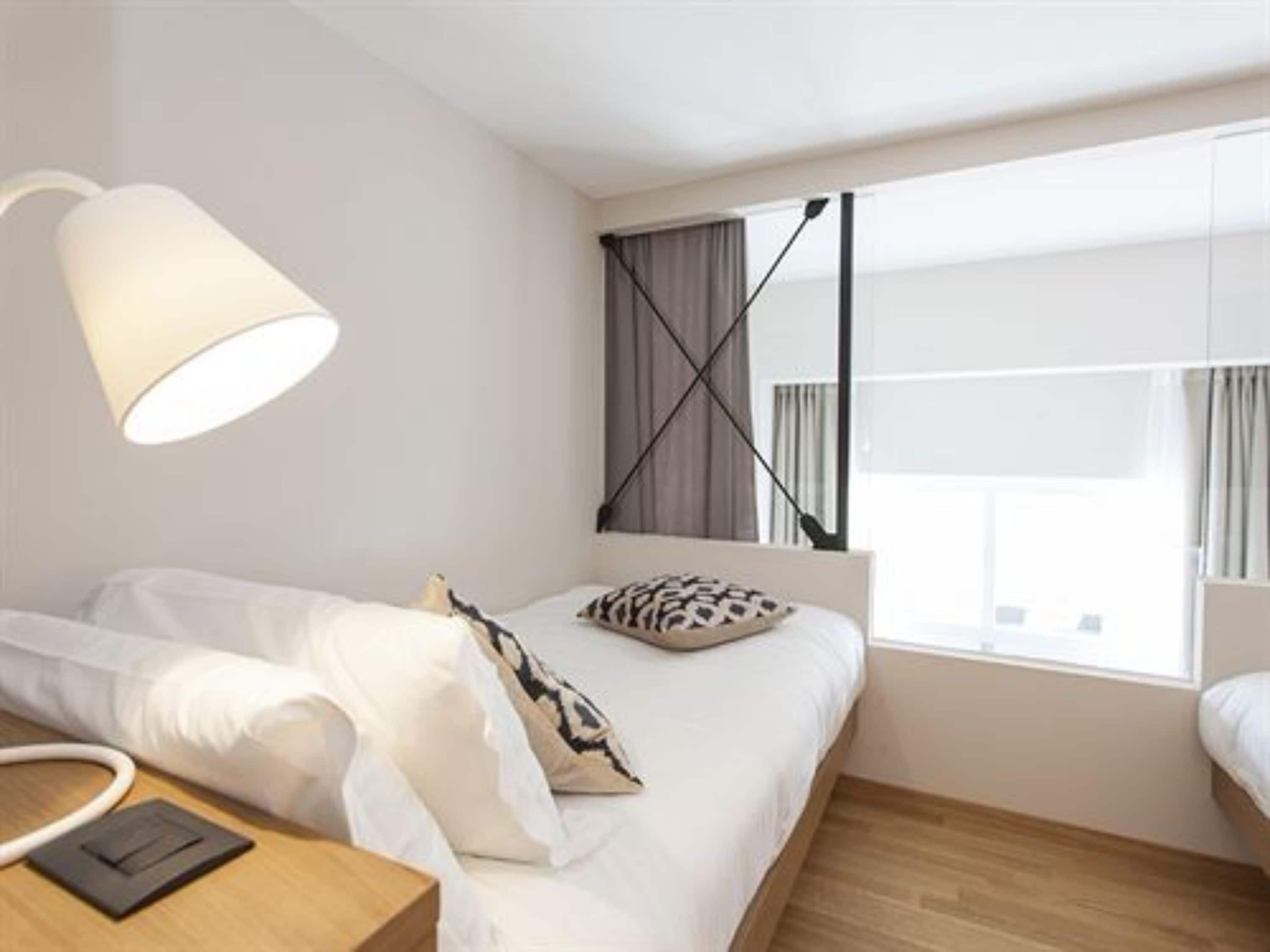 A modern hotel room with a bed, a bedside table, a lamp, and a window with curtains.