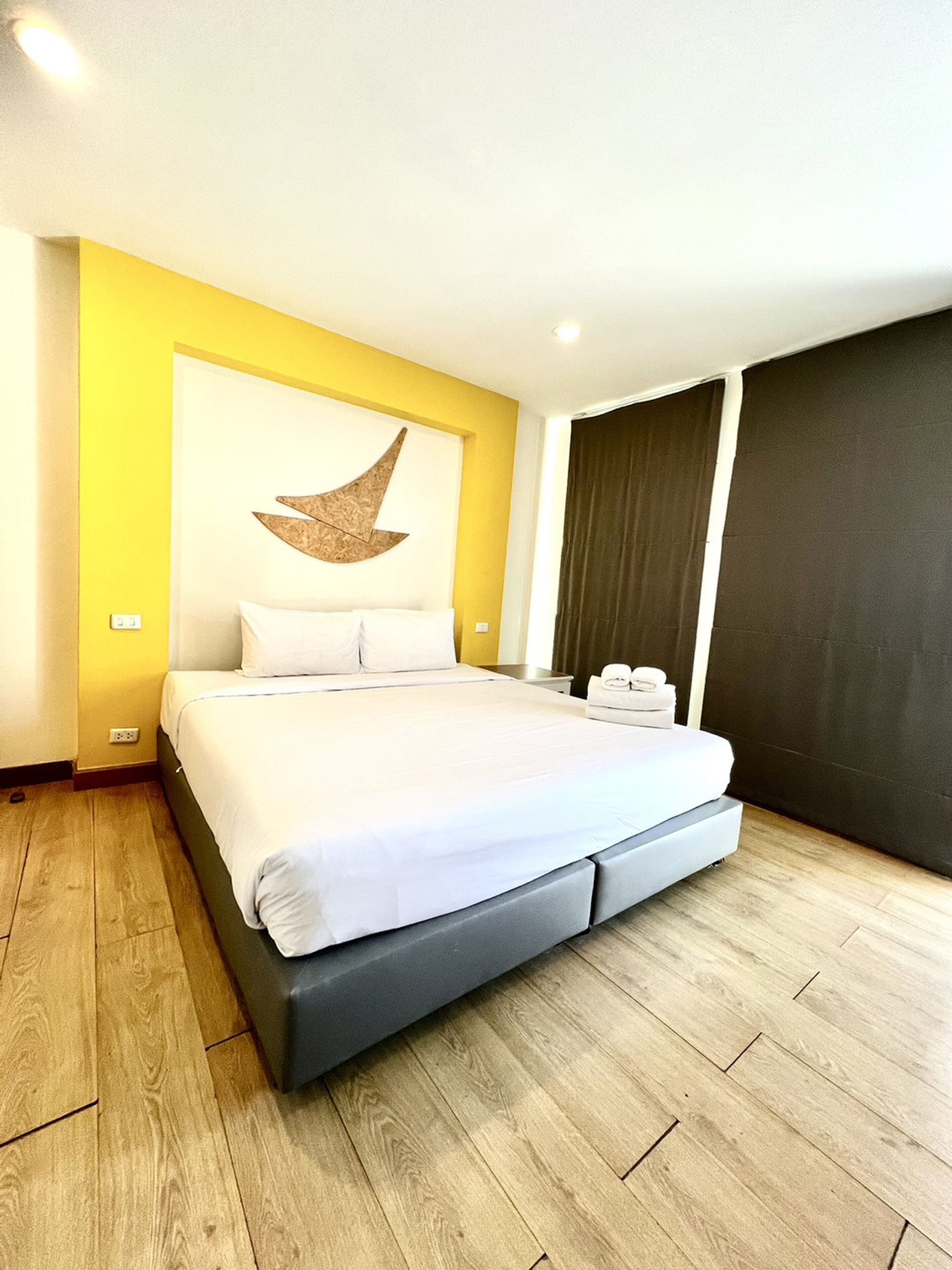 A modern hotel room with a large bed, wooden flooring, a yellow accent wall, and a large window with curtains.