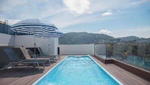 Una piscina en la azotea con sillones de descanso y sombrillas, rodeada de barandillas de cristal y vistas a una colina arbolada.