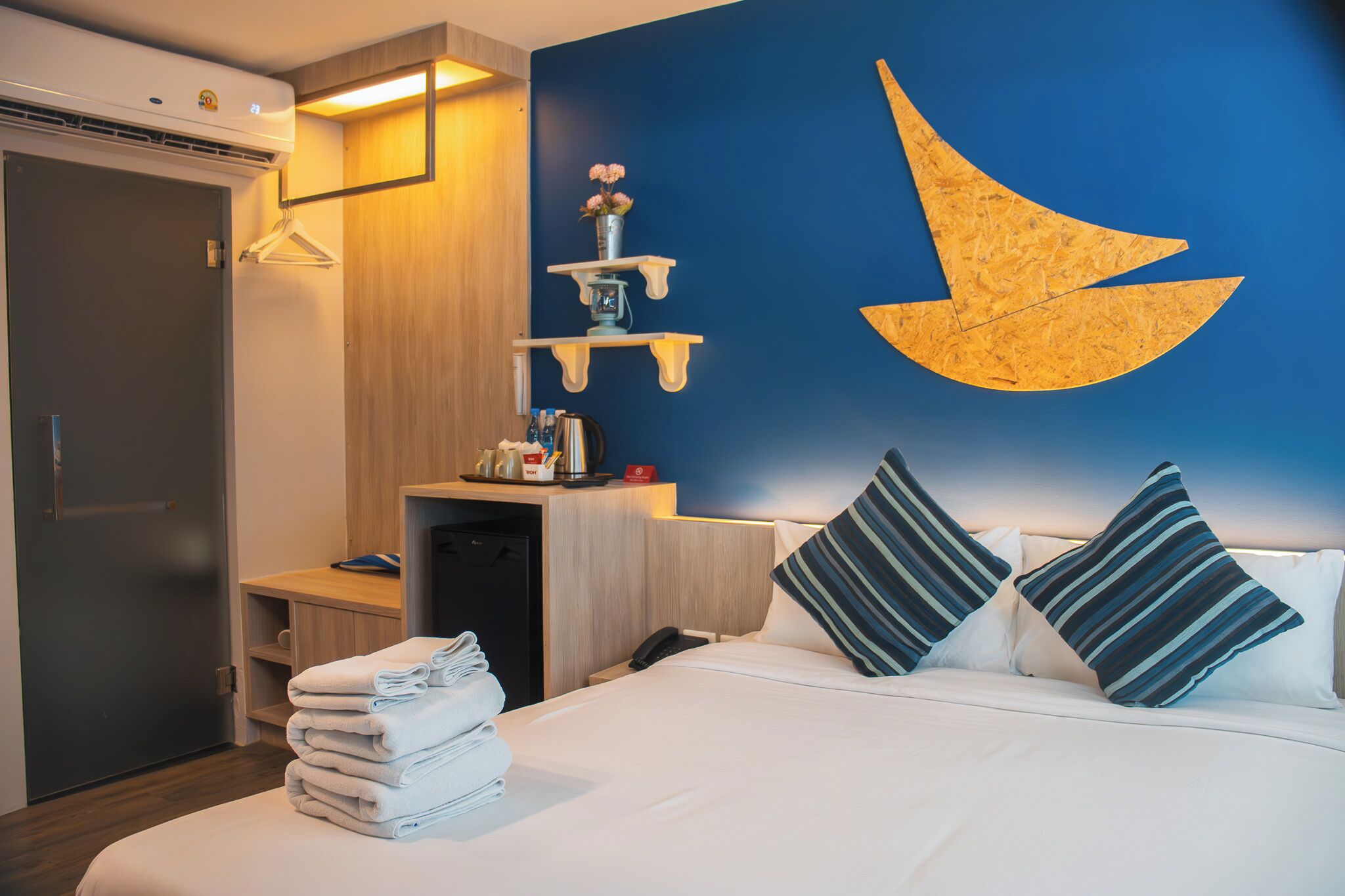 A hotel room with a bed, a desk, a wardrobe, and a wall-mounted shelf with a vase of flowers.