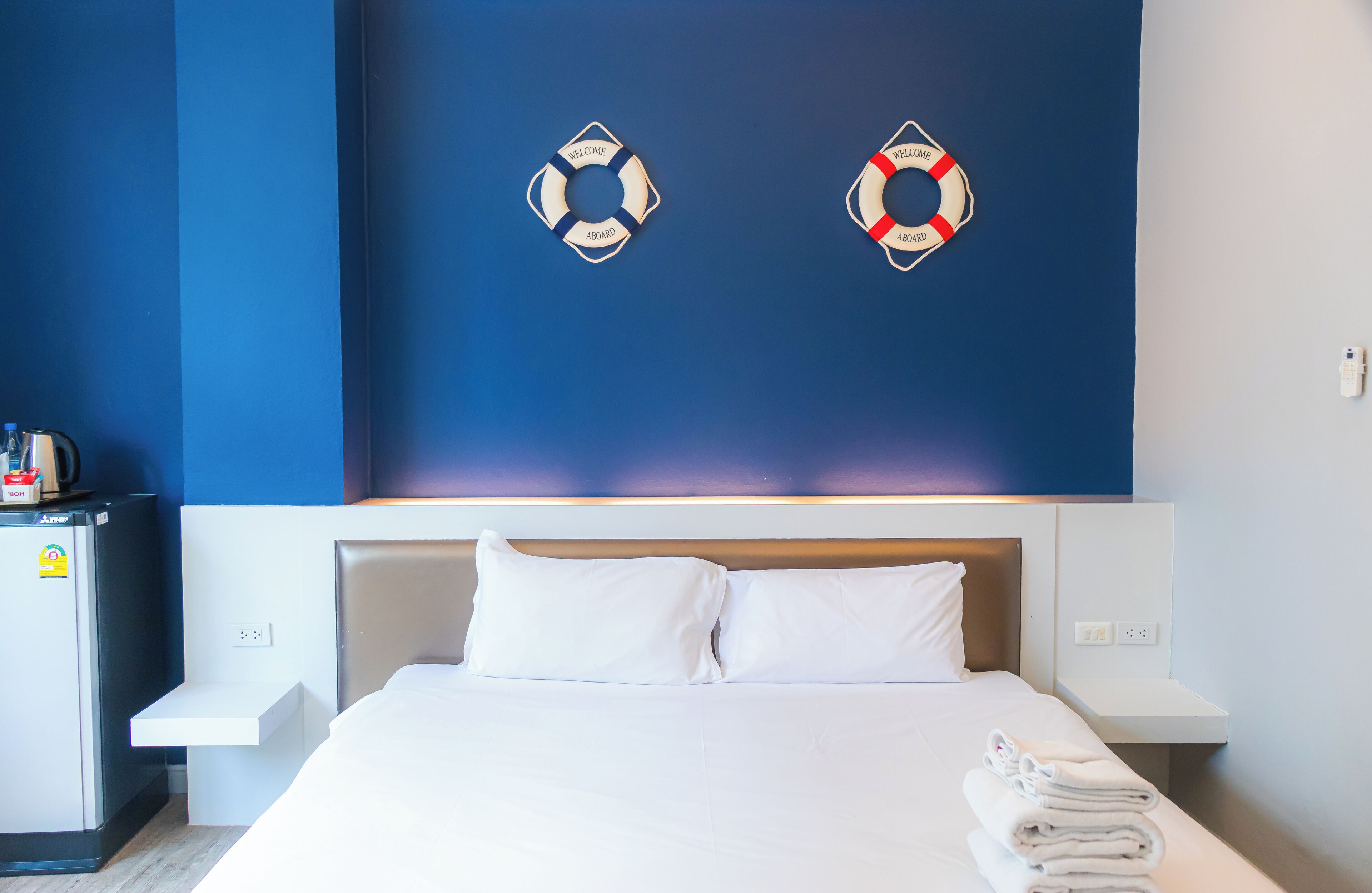 A hotel room with a bed, white pillows, a small bedside table, a nightstand, and a wall-mounted life preserver decoration.