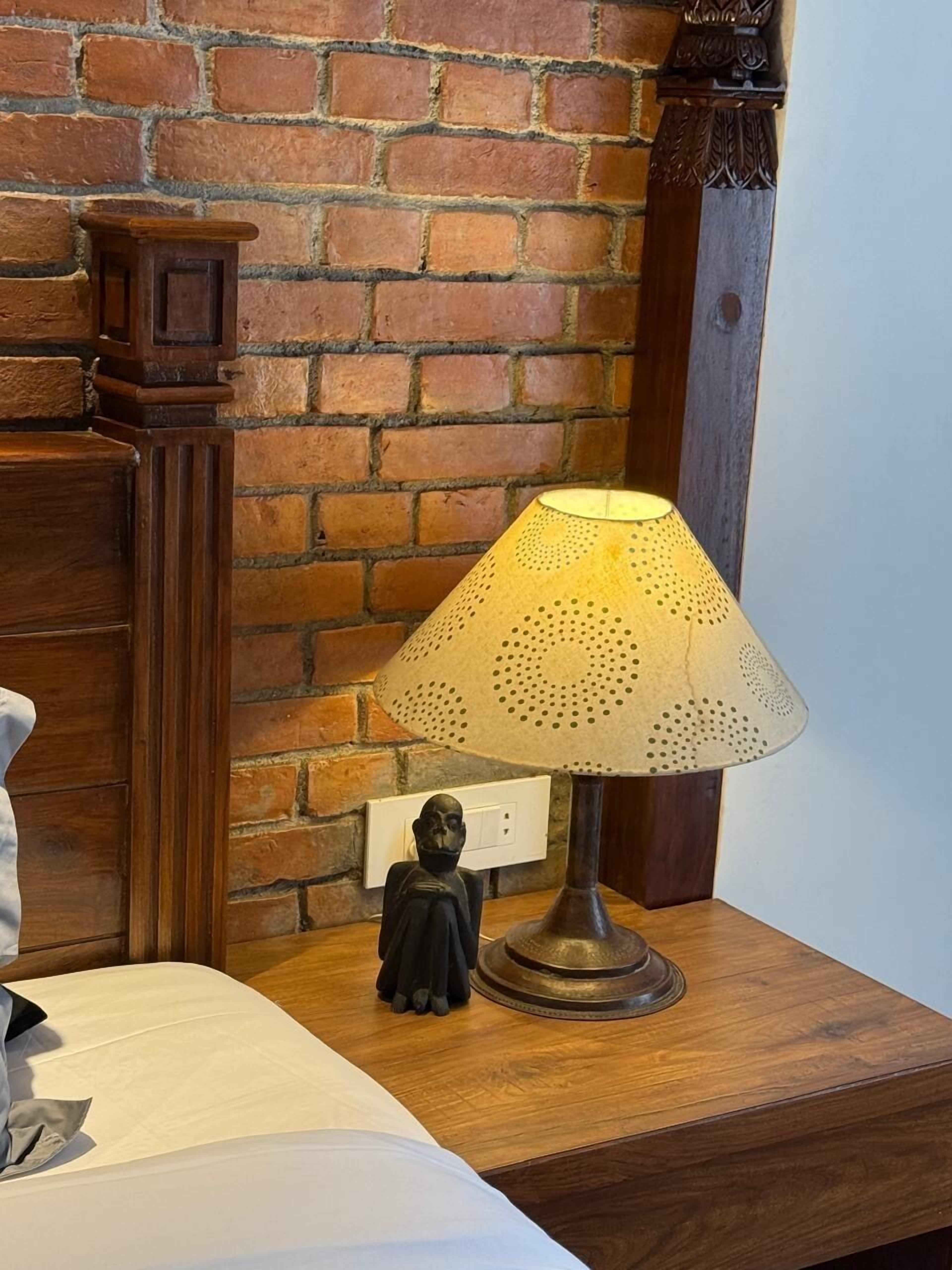 Una mesita de noche de madera con una lámpara, un pequeño mueble auxiliar y una toma de corriente.
