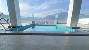 Una piscina en la azotea con baldosas azules cristalinas, rodeada de pilares blancos, y que ofrece vistas a un paisaje urbano y montañas a lo lejos.