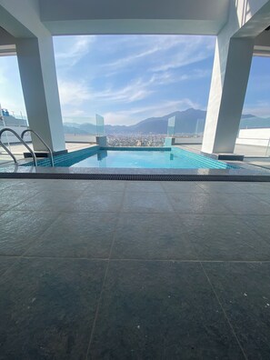 Una piscina en la azotea con baldosas azules cristalinas, rodeada de pilares blancos, y que ofrece vistas a un paisaje urbano y montañas a lo lejos.