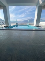 A rooftop pool with clear blue tiles, surrounded by white pillars, and offering a view of a cityscape and mountains in the distance.