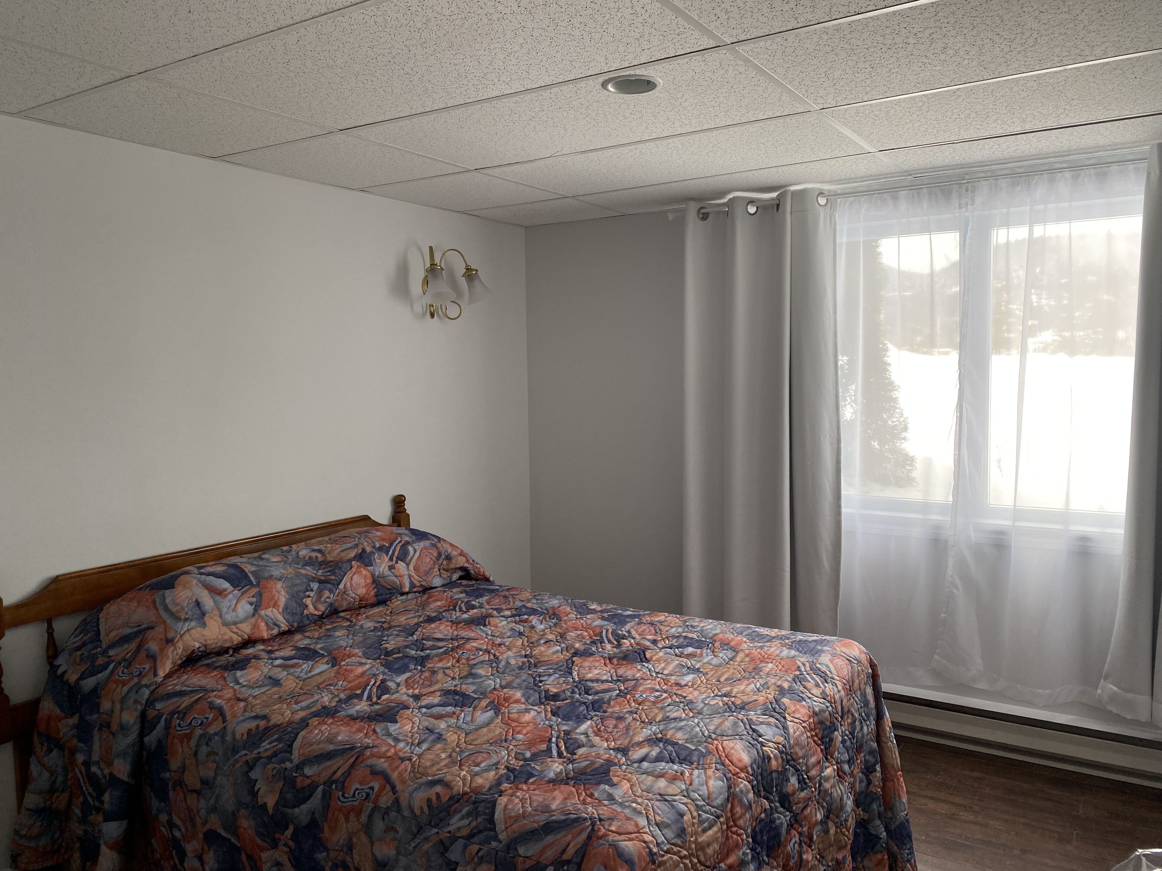 Ein Schlafzimmer mit einem hölzernen Bett, einer gemusterten Tagesdecke, einer weißen Wand, einer Decke mit punktförmigem Muster und einem Fenster mit durchsichtigen Vorhängen.