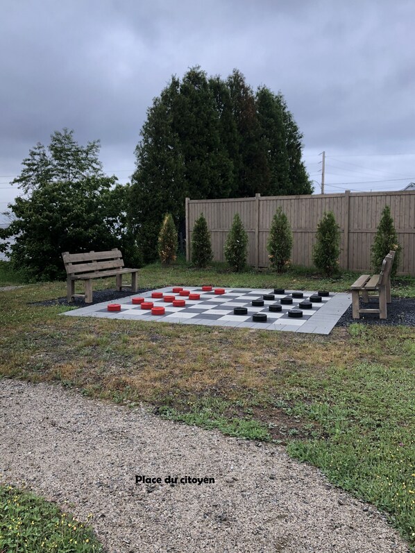 A checkered outdoor game area with benches, a wooden fence, and trees in the background. - À Fleur de Baie (Baie Sainte-Catherine)