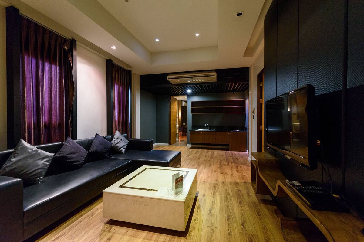 Uma sala de estar moderna com um sofá de cor escura, uma mesa de centro de vidro e uma televisão de tela plana.