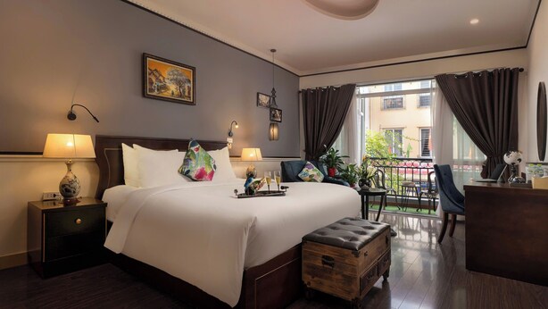 Habitación de hotel con una cama grande, lámparas de noche, un cofre de madera, un escritorio y un balcón con plantas.