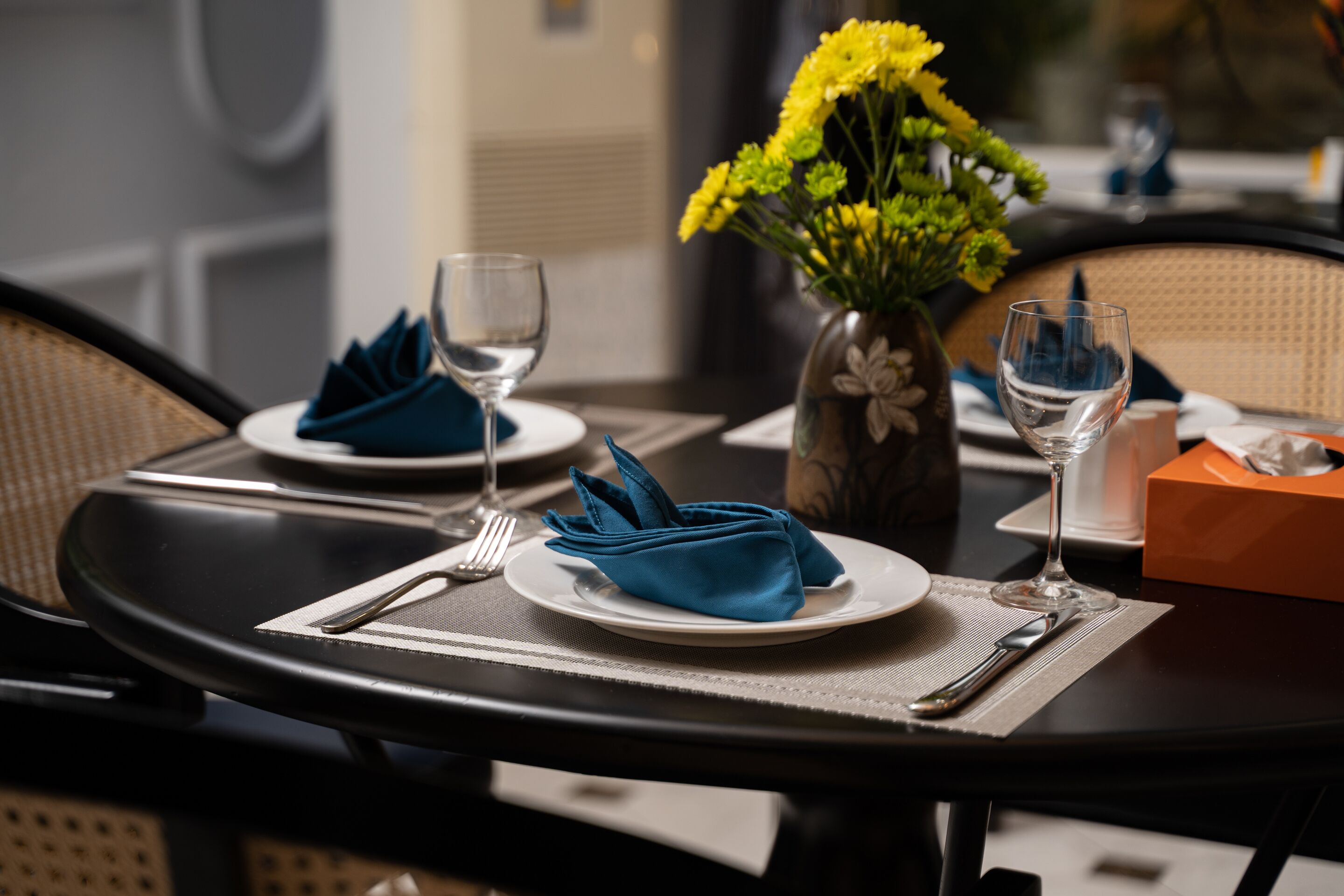 A dining table set with plates, folded napkins, cutlery, and a vase with yellow flowers.