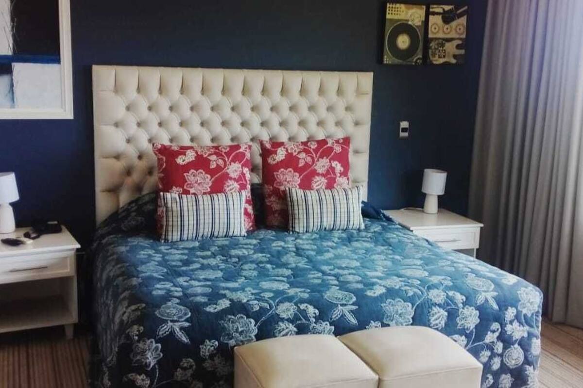 A bedroom with a tufted headboard, a floral bedspread, two bedside tables with lamps, and a window with curtains.