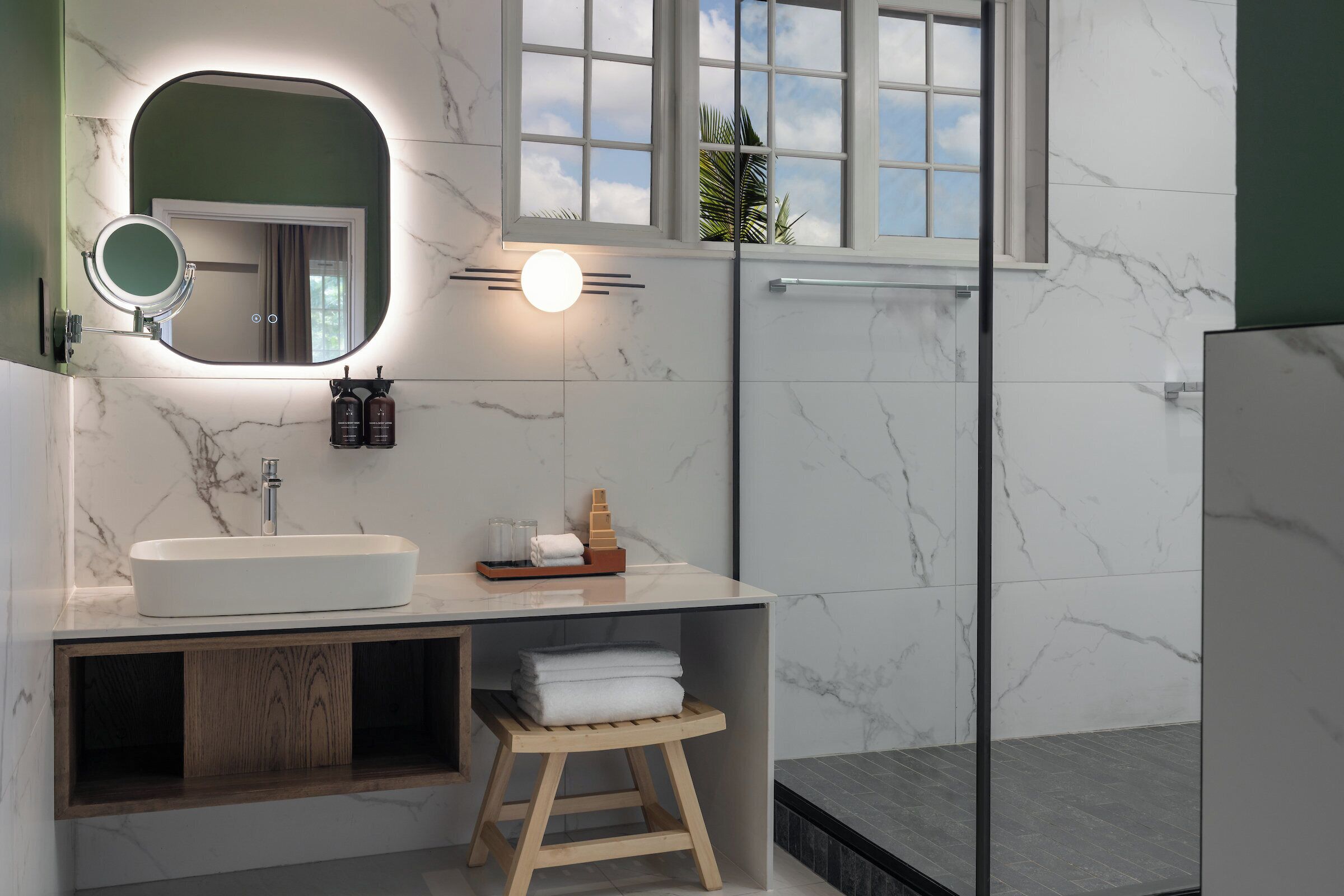 Un baño moderno con un espejo grande, una lámpara redonda, un lavamanos y una ventana con vistas al cielo.