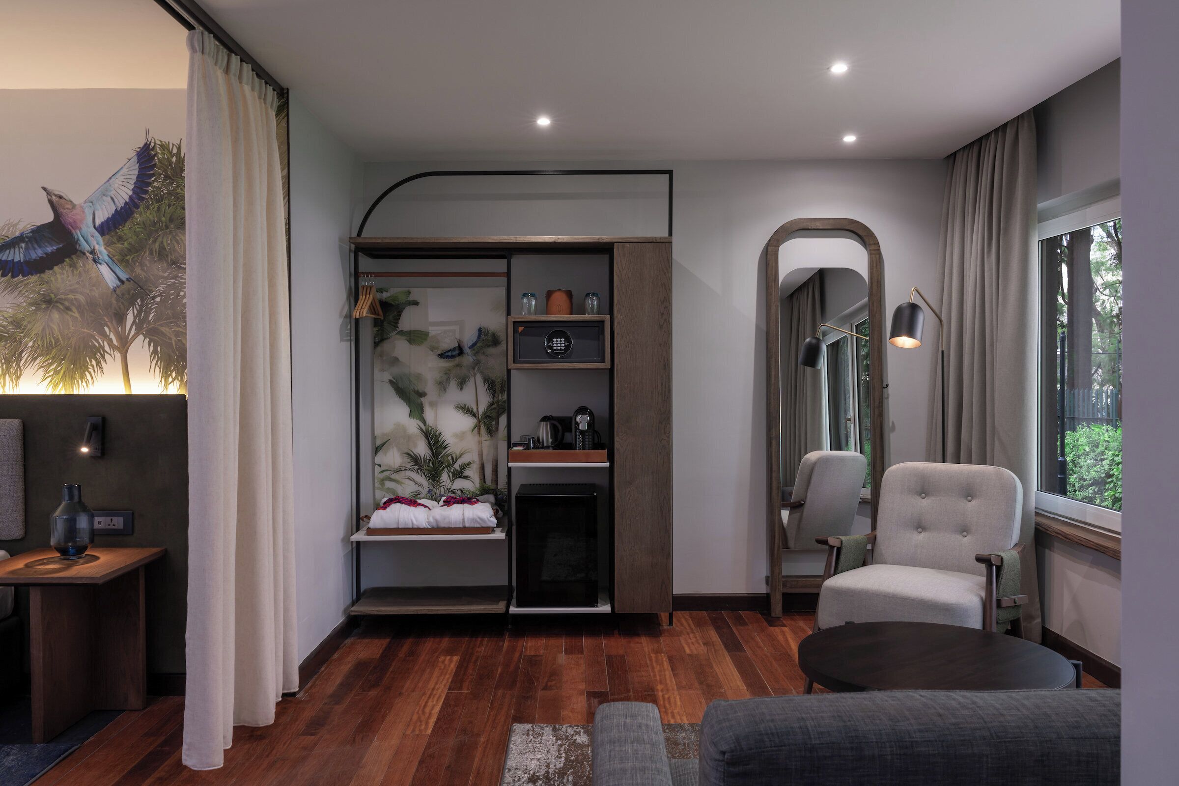 Una sala de estar moderna con piso de madera, un sofá gris, una pequeña mesa redonda, un sillón gris, una estantería empotrada con objetos decorativos, una chimenea y un ventanal con cortinas.