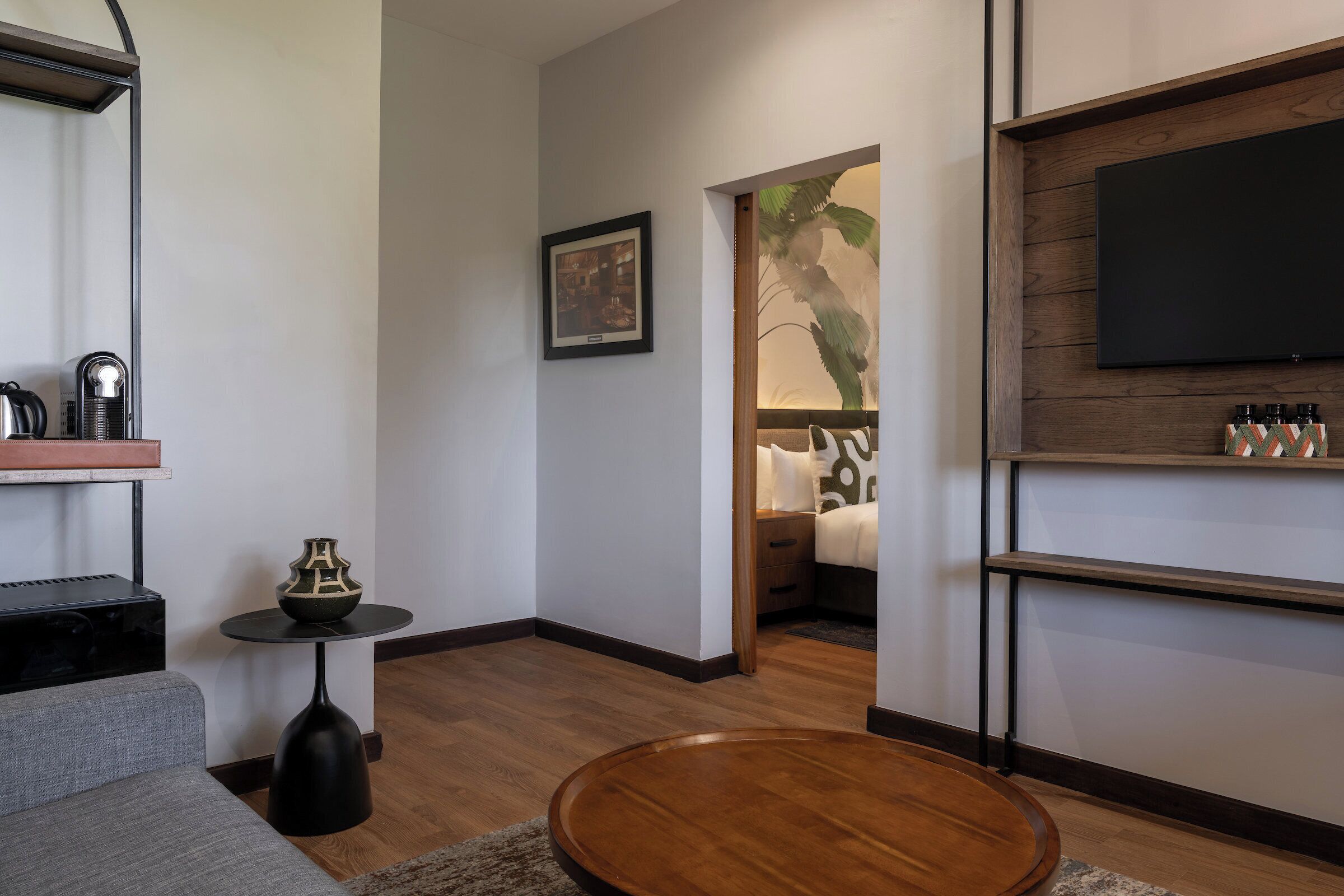 Una sala de estar moderna con un sofá gris, una mesa de centro redonda de madera, una mesita auxiliar negra con un florero, un cuadro en la pared, un televisor de pantalla plana montado en una pared de madera y un estante con objetos decorativos.