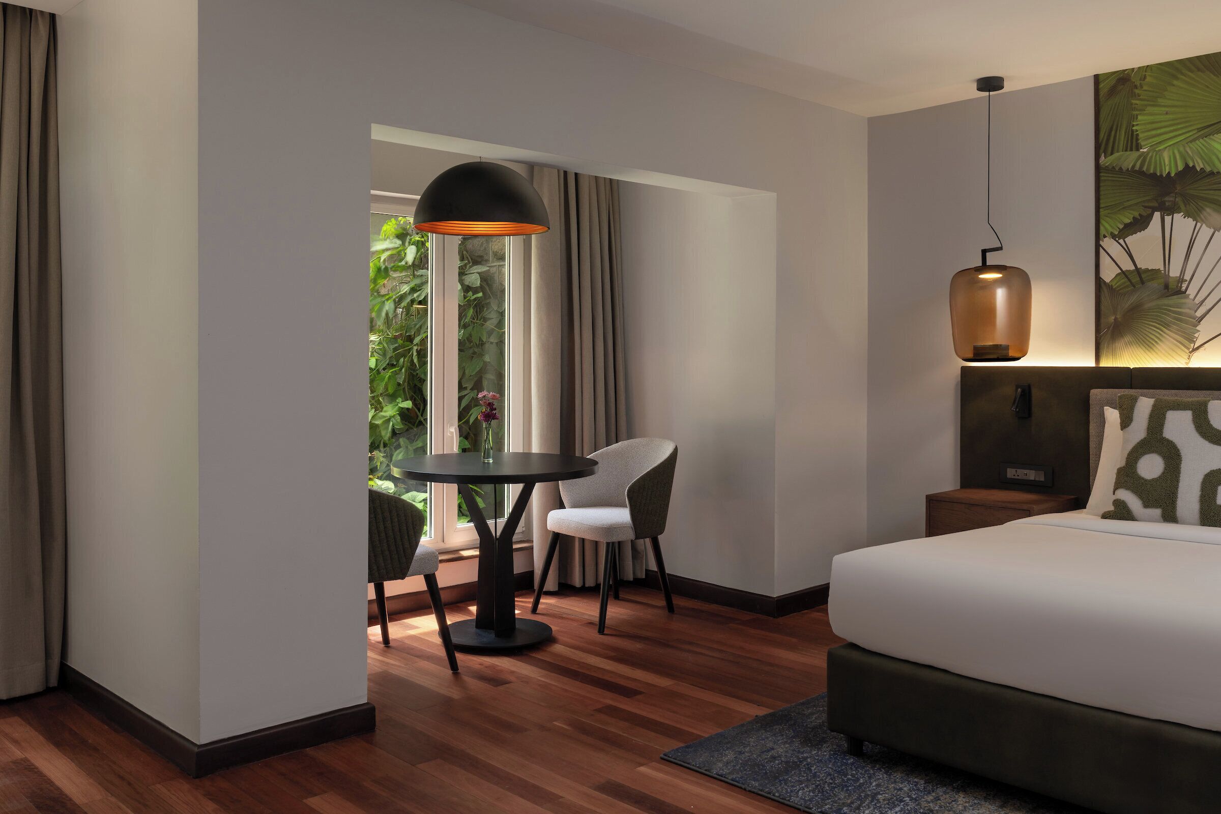 A hotel room with a bed, a round table with two chairs, a large window with curtains, and a hanging pendant light.