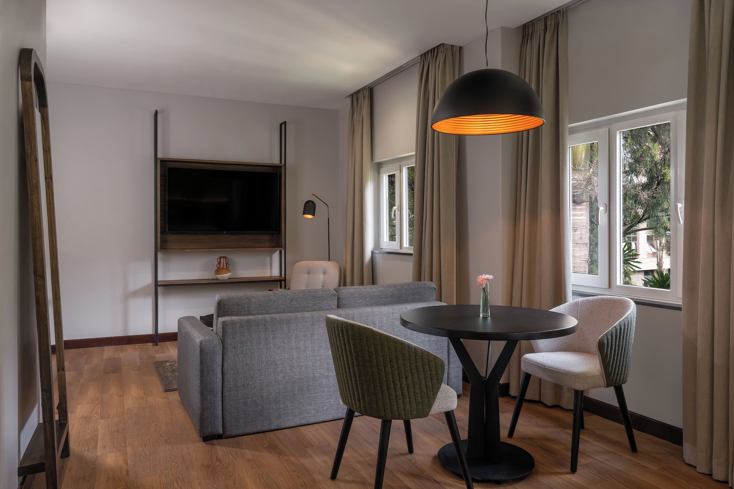Ein modernes Wohnzimmer mit einem grauen Sofa, zwei Sesseln, einem runden Tisch, einem an der Wand montierten Fernseher und einem Fenster mit Vorhängen.