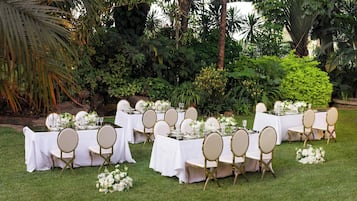 An outdoor wedding setup with tables covered in white tablecloths, gold chairs, and floral centerpieces.