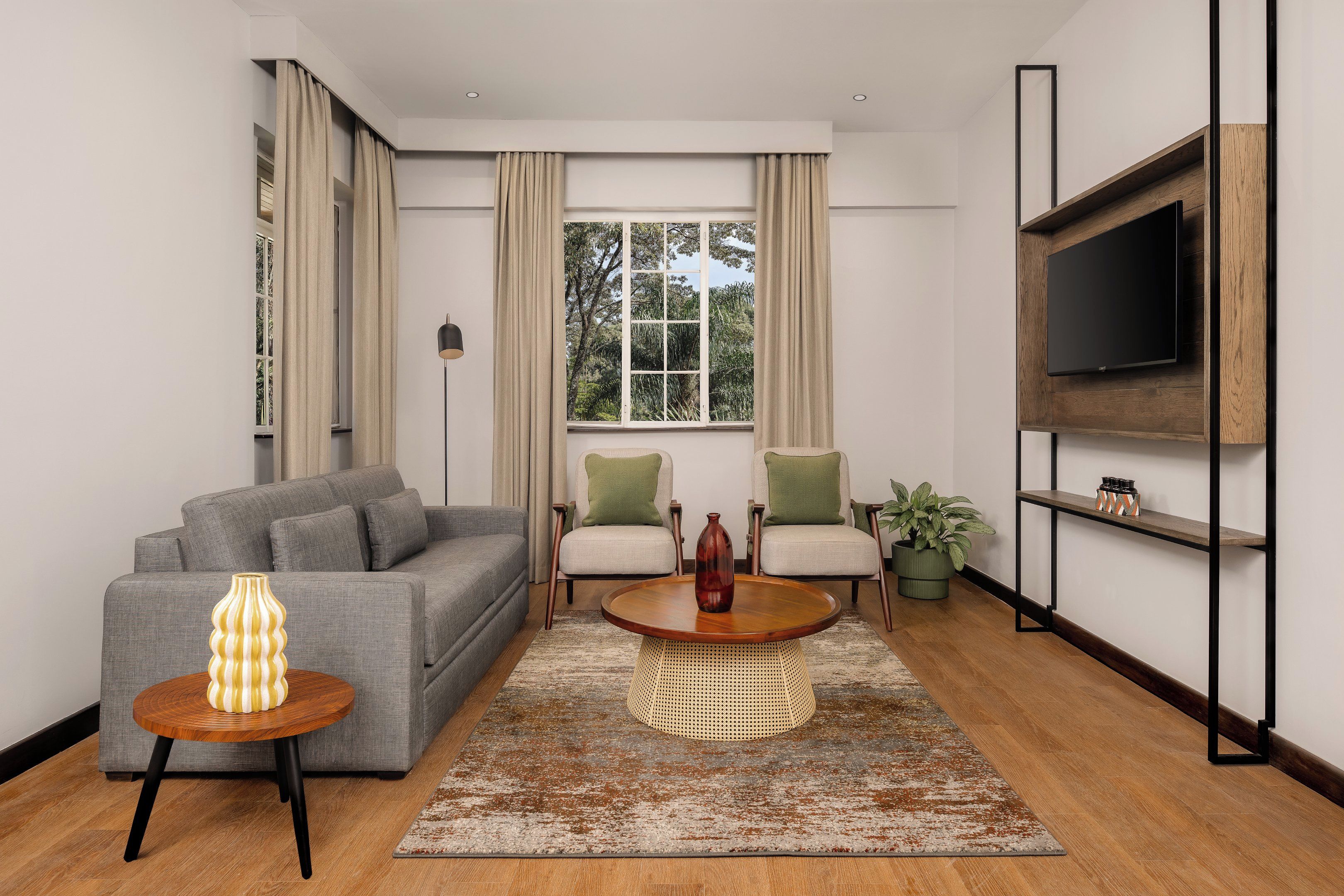 A modern living room with a grey sofa, two armchairs, a coffee table, a TV mounted on a wooden shelf, and a large window with curtains.