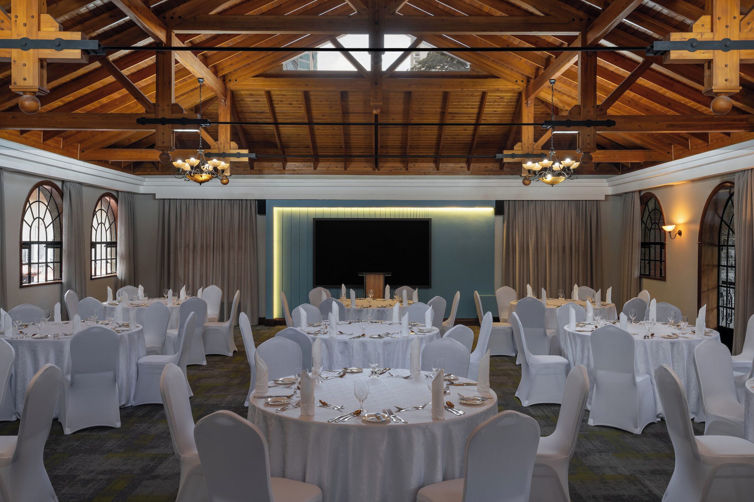 A banquet hall with round tables set for dining, white chairs, chandeliers, and a stage with curtains.