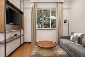A modern living room with a flat-screen TV, a round wooden coffee table, a grey sofa, and a large window with curtains.