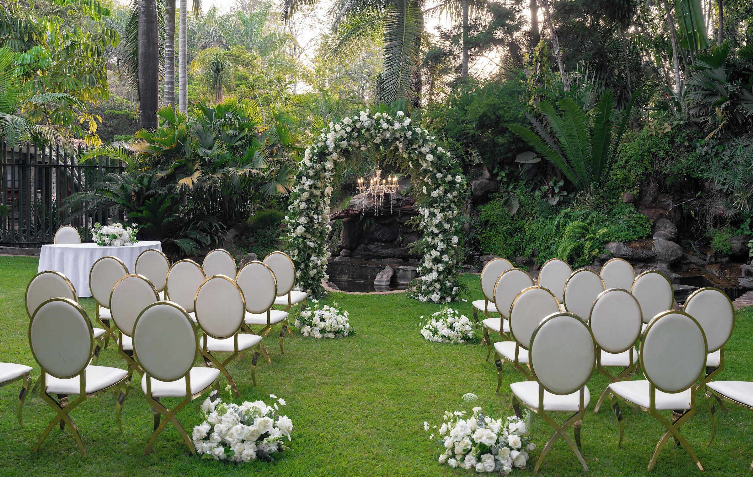 An outdoor wedding venue with a floral arch, white chairs, and a small waterfall.