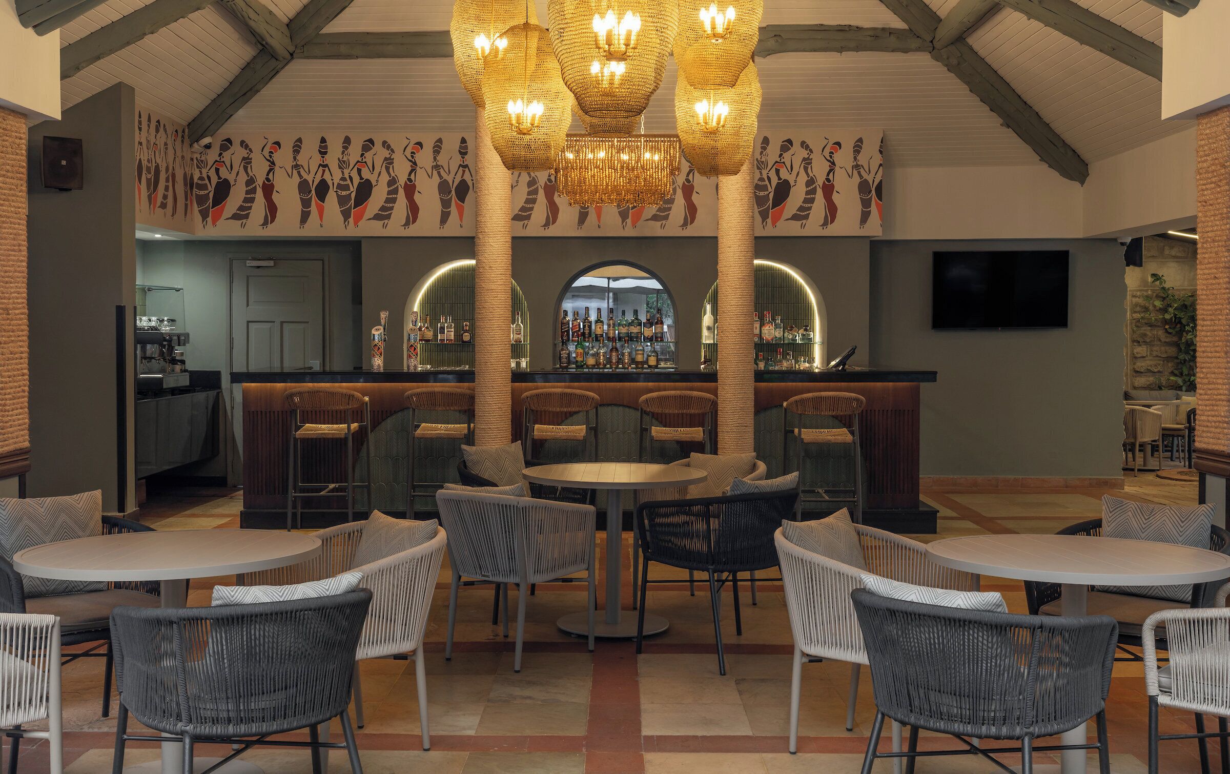 A bar area with wicker chairs and tables, a bar counter with bottles, and a decorative ceiling.