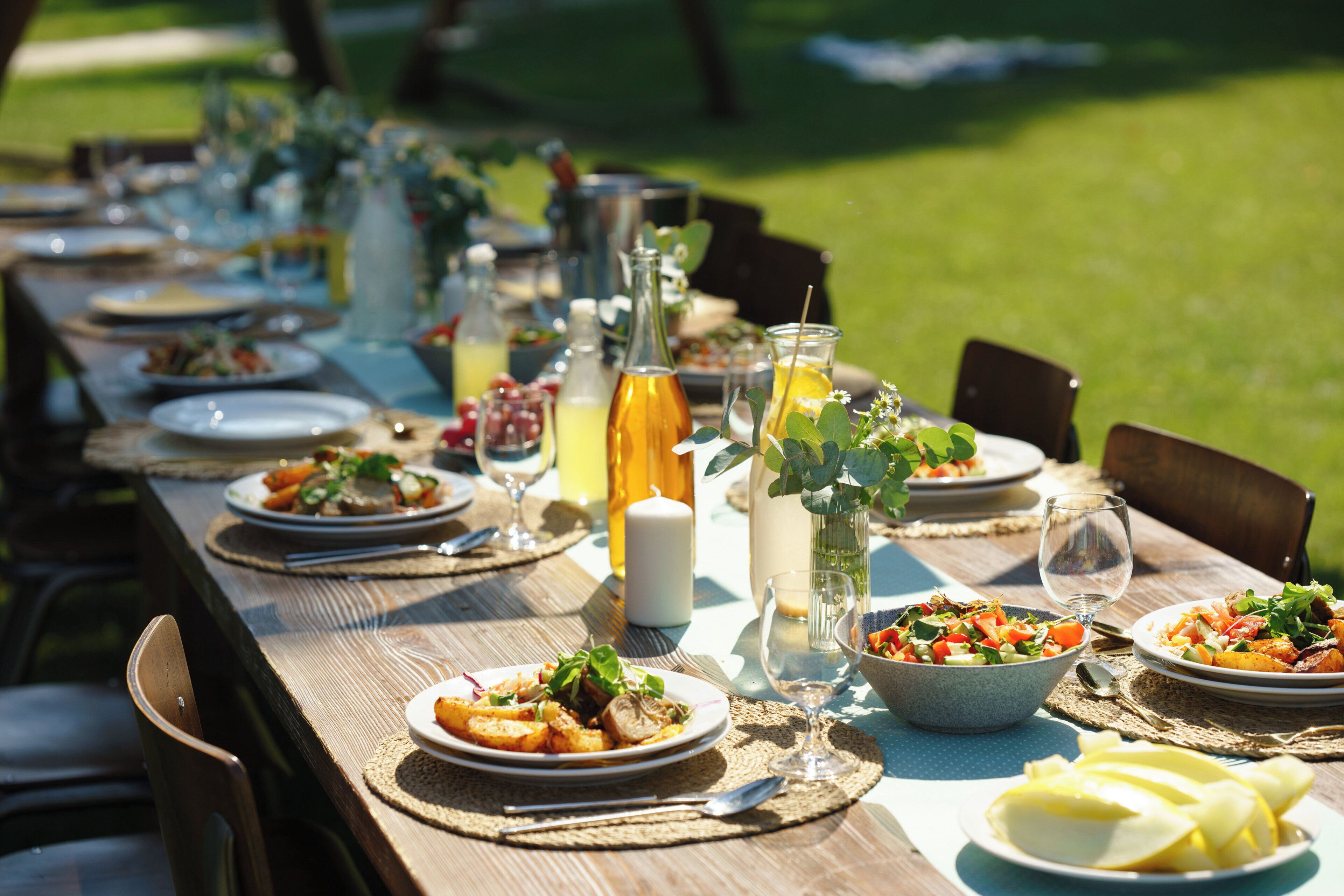 A long wooden table set for a meal outdoors with plates of food, glasses, and bottles.