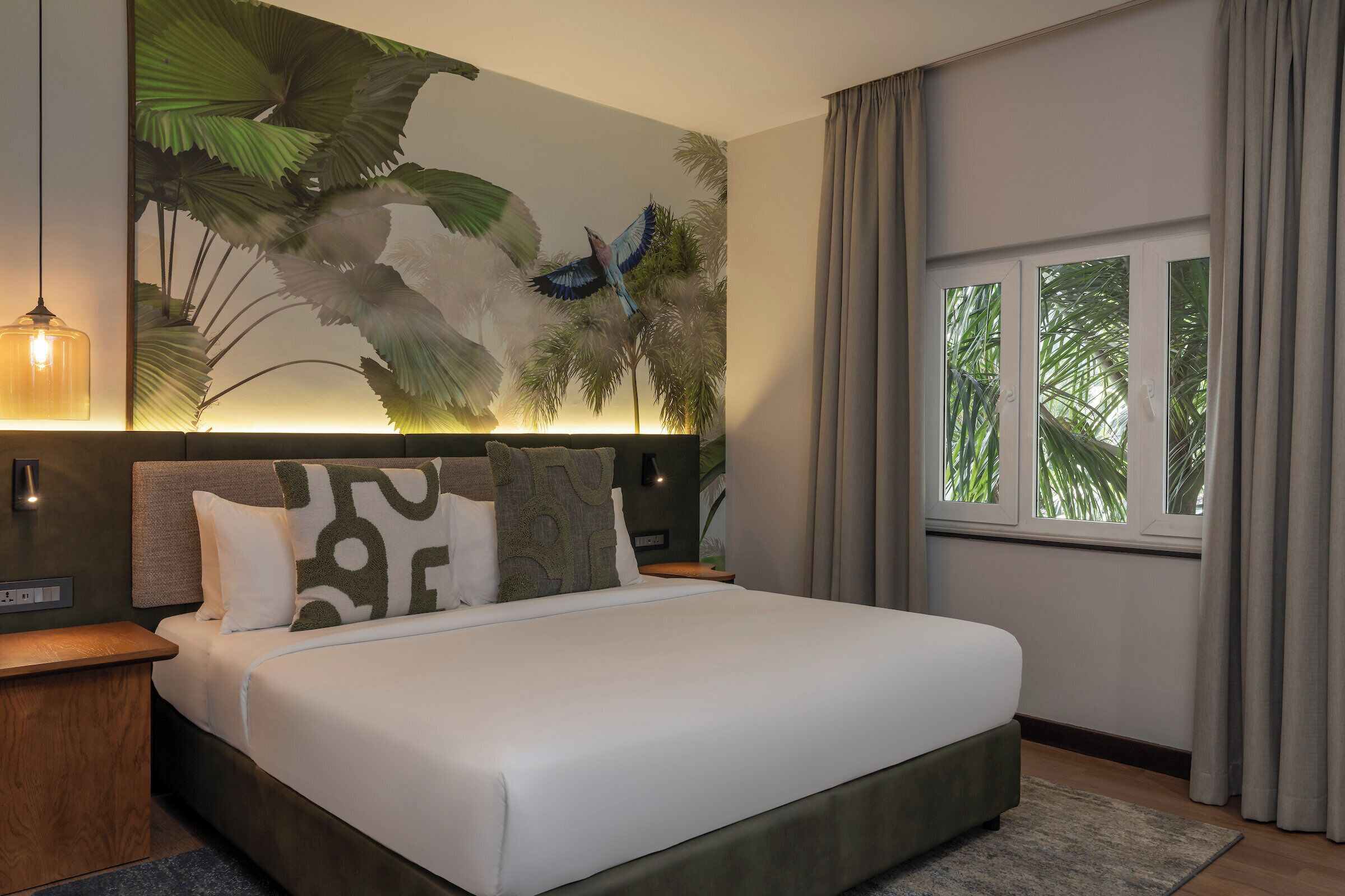 A modern hotel room with a large bed, a nightstand, a wall-mounted lamp, and a window with curtains.