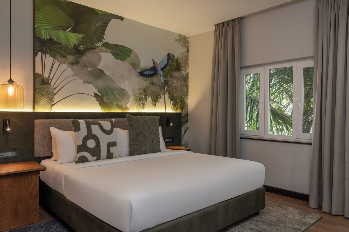 Una habitación de hotel moderna con una cama grande, una mesita de noche, una lámpara de pared y un ventanal con cortinas.