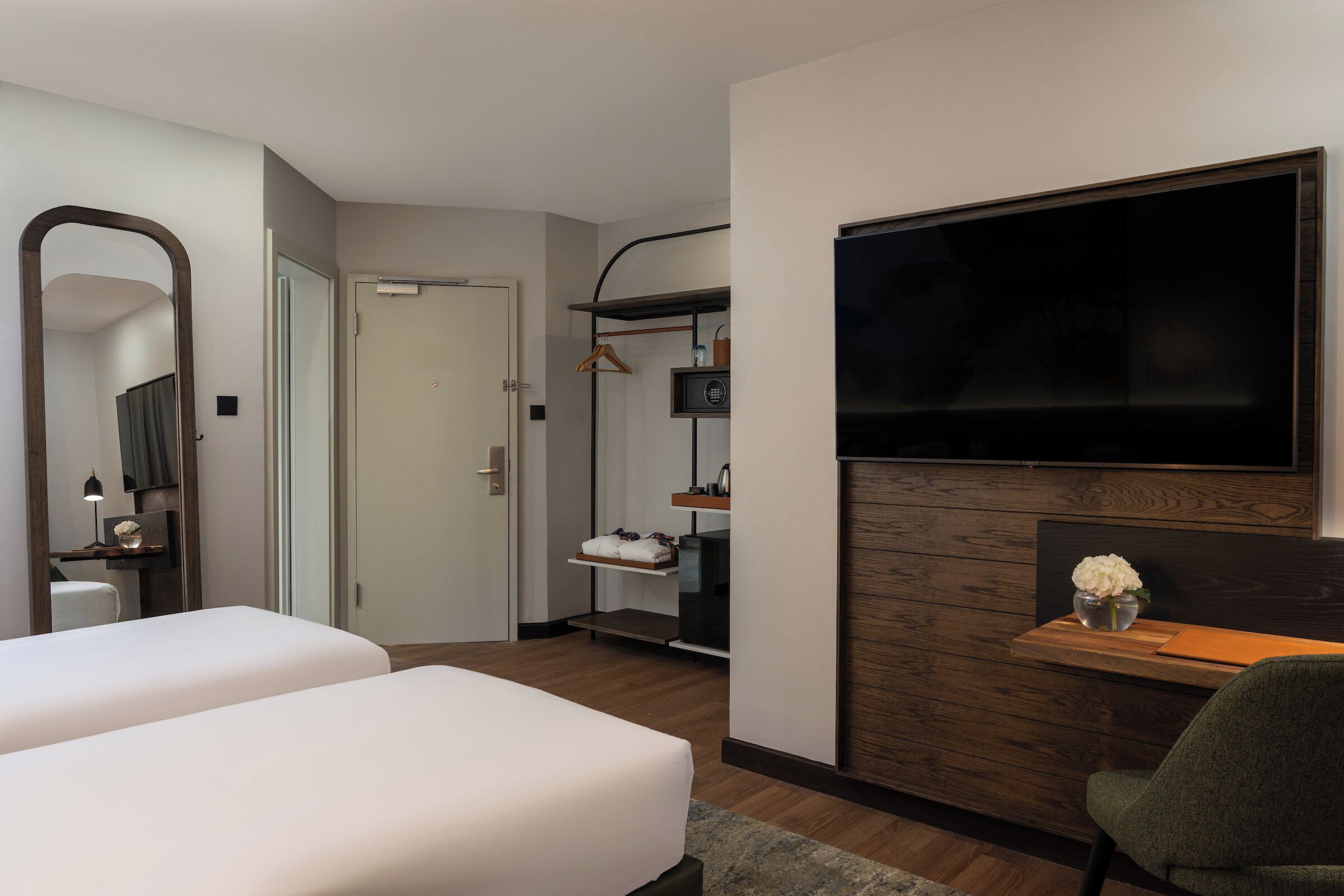 Habitación de hotel con dos camas, televisión, escritorio con silla, espejo, armario y estante con artículos de tocador.