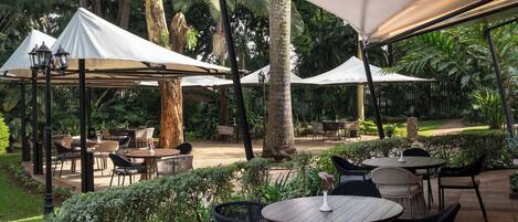 An outdoor patio area with round tables and chairs, covered by white canopies, surrounded by lush greenery and palm trees.