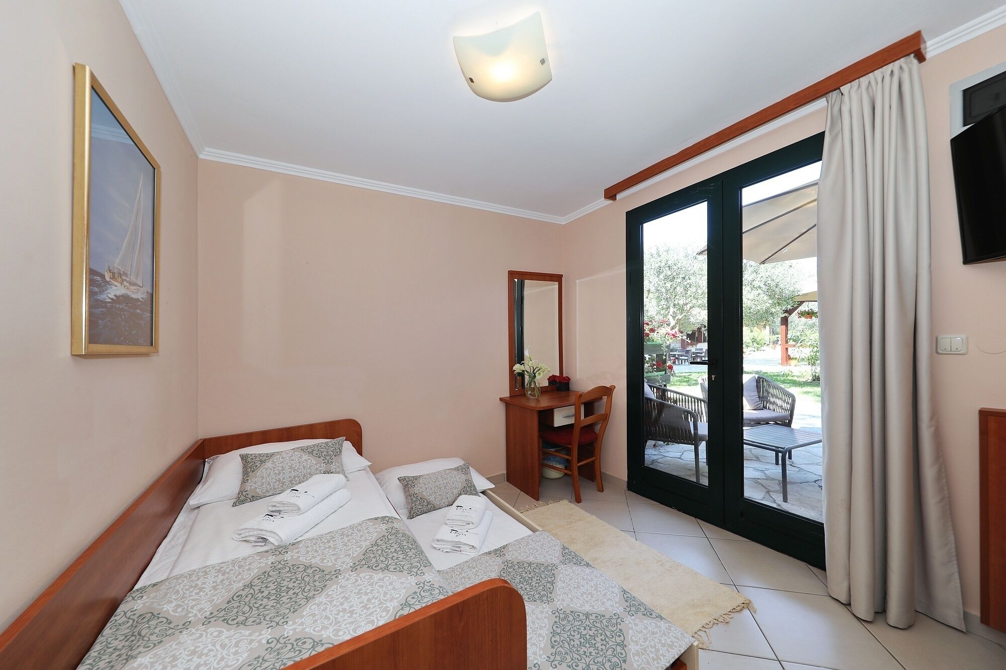 Un dormitorio con cama, escritorio, silla, espejo y una puerta corrediza de vidrio que da a un patio al aire libre.