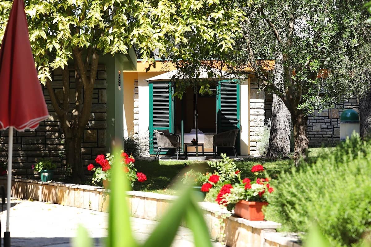 Un bâtiment en pierre avec des volets verts, un petit porche avec deux chaises et des fleurs rouges dans des jardinières.