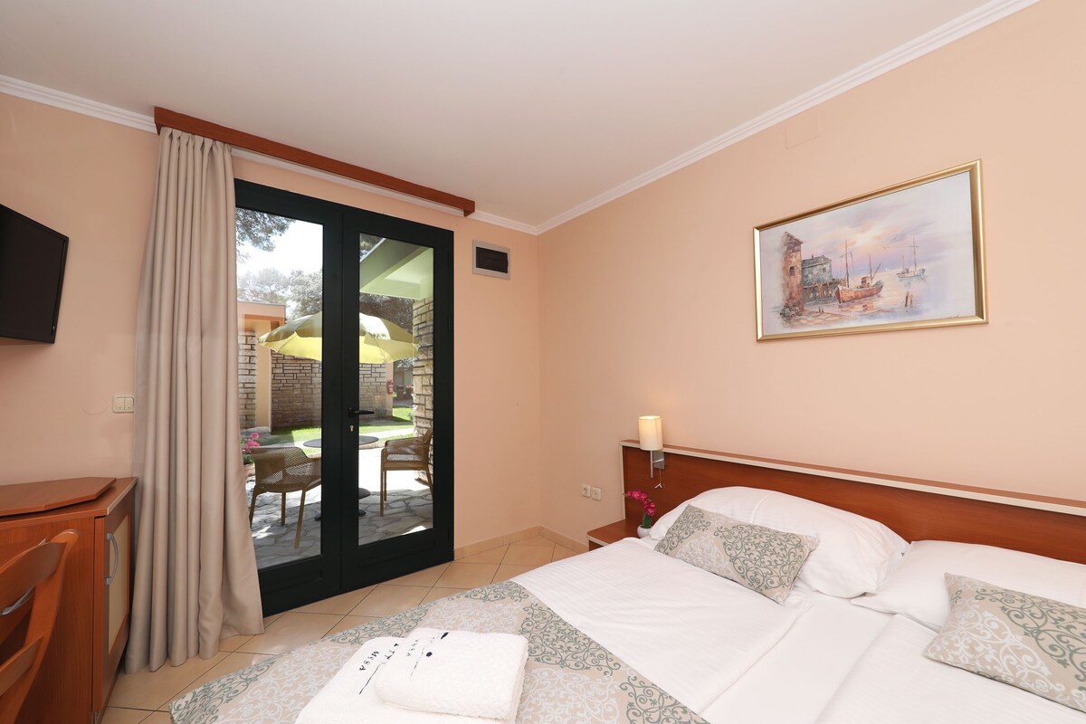Une chambre d’hôtel avec un lit, un bureau, une chaise, un tableau et une vue sur un espace extérieur accessible par une porte vitrée, cet espace étant aménagé avec une table et des chaises.
