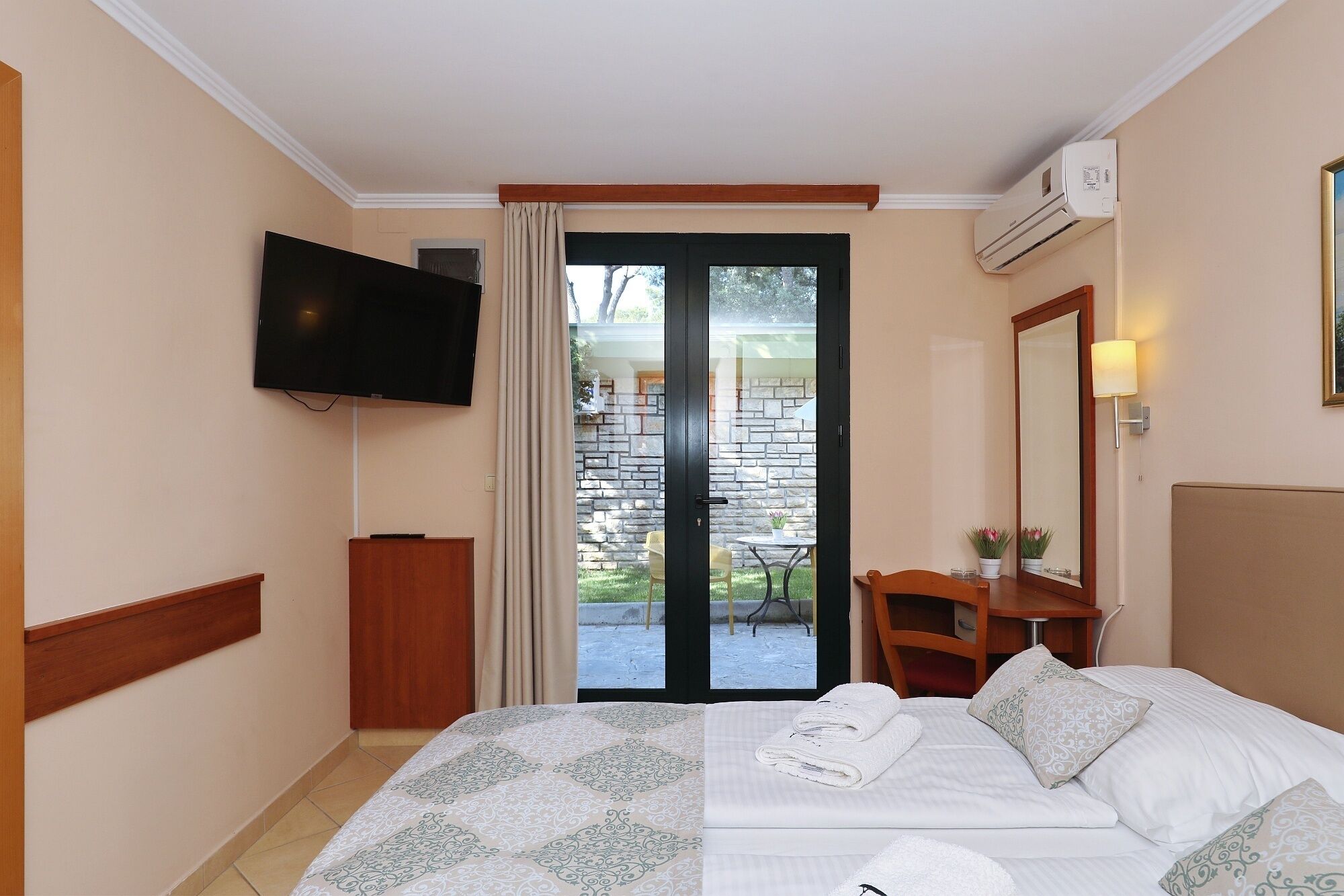 A hotel room with a bed, a TV, a desk with a chair, a mirror, and a view of the outside.