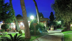 A well-lit pathway leading to a building with a tiled roof and large windows, surrounded by lush greenery and trees. - The Mediterranean Village San Antonio (Biograd na Moru)