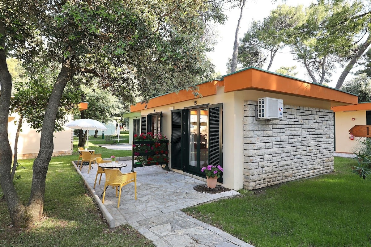 Un petit bâtiment d’un seul étage, doté d’une façade en pierre et d’un toit orange. Il comprend un patio avec des sièges extérieurs et un pot de fleurs.