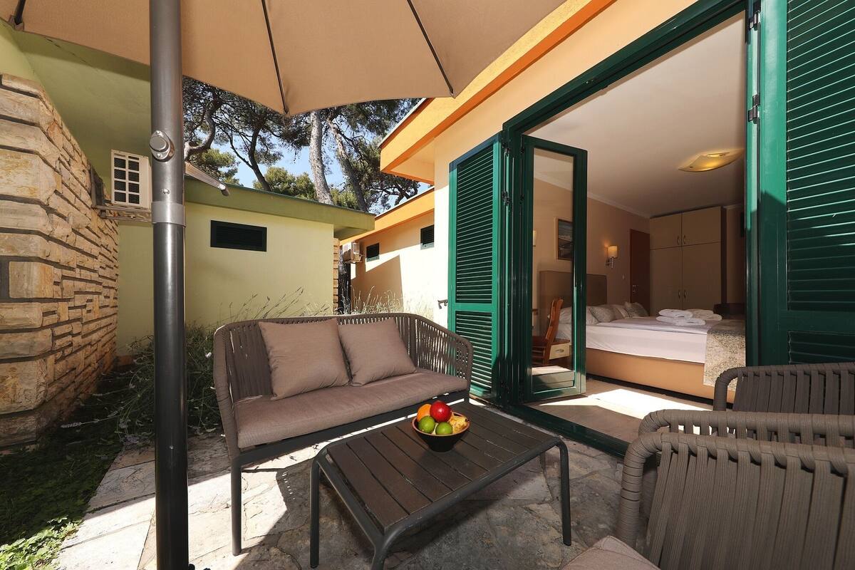 Une chambre d’hôtel avec un lit, un canapé et une table sur laquelle se trouvent des fruits. Il y a un balcon avec vue sur les arbres et un mur en pierre.