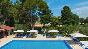 A swimming pool with blue water, surrounded by lounge chairs and umbrellas, with trees and buildings in the background. - The Mediterranean Village San Antonio (Biograd na Moru)