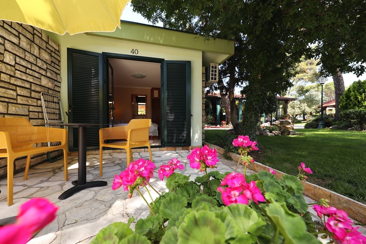 L’entrée d’un hôtel, avec un mur en pierre, le numéro 40, et un patio abrité agrémenté de chaises jaunes et de fleurs roses.