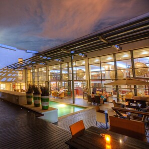 Un espacio al aire libre con mesas y sillas, una piscina y un edificio con paredes de vidrio y luz interior.